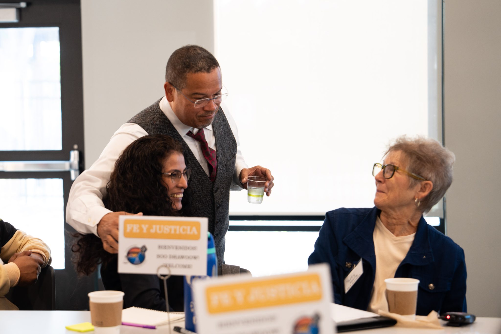 Three people laughing in a conference room. Placards on the tables say "Fe y Justicia"