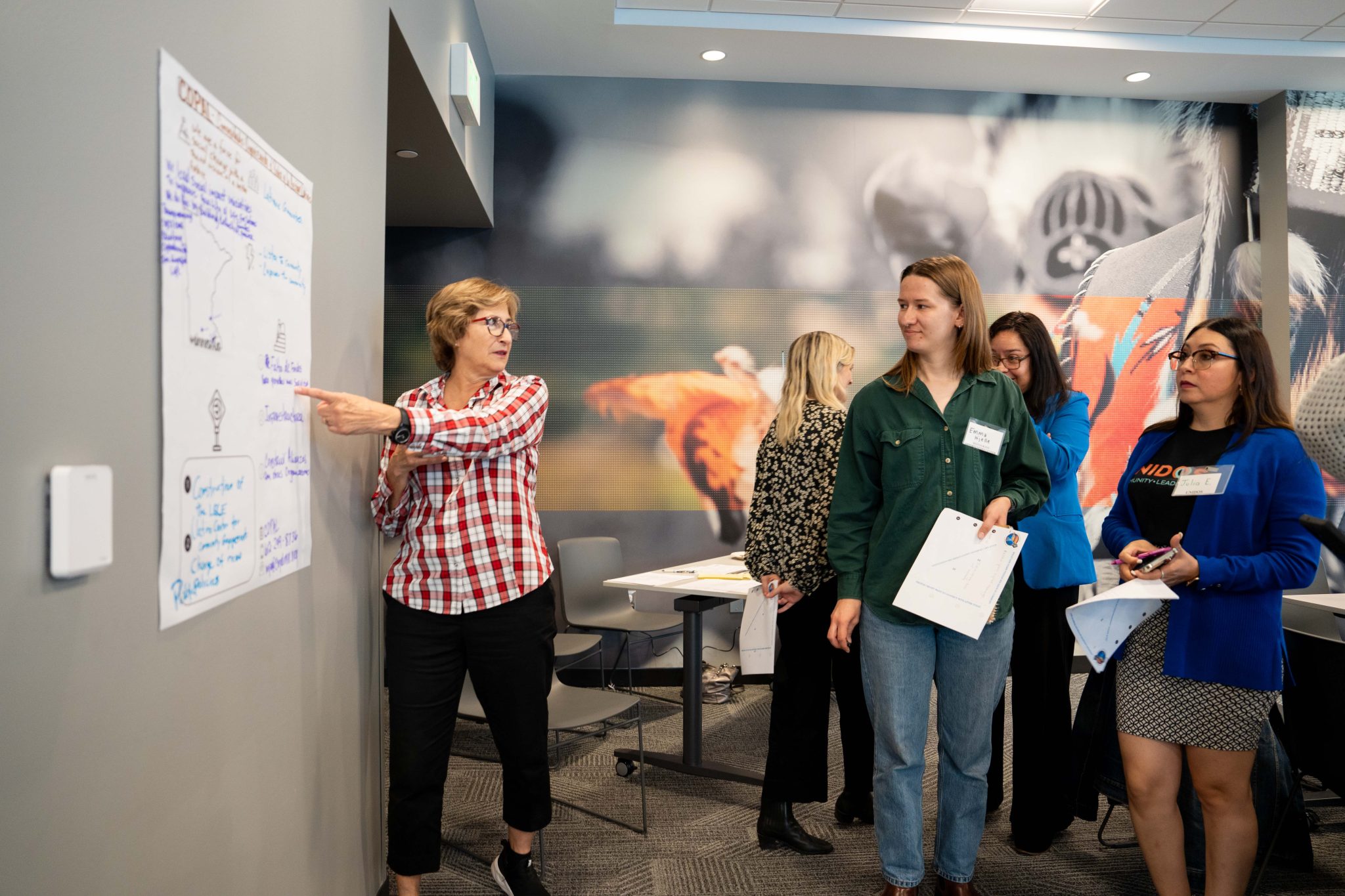 A woman points to show four other women a chart with hand written markings on the wall