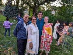 A family of four poses outdoors. More family members are playing in the background.