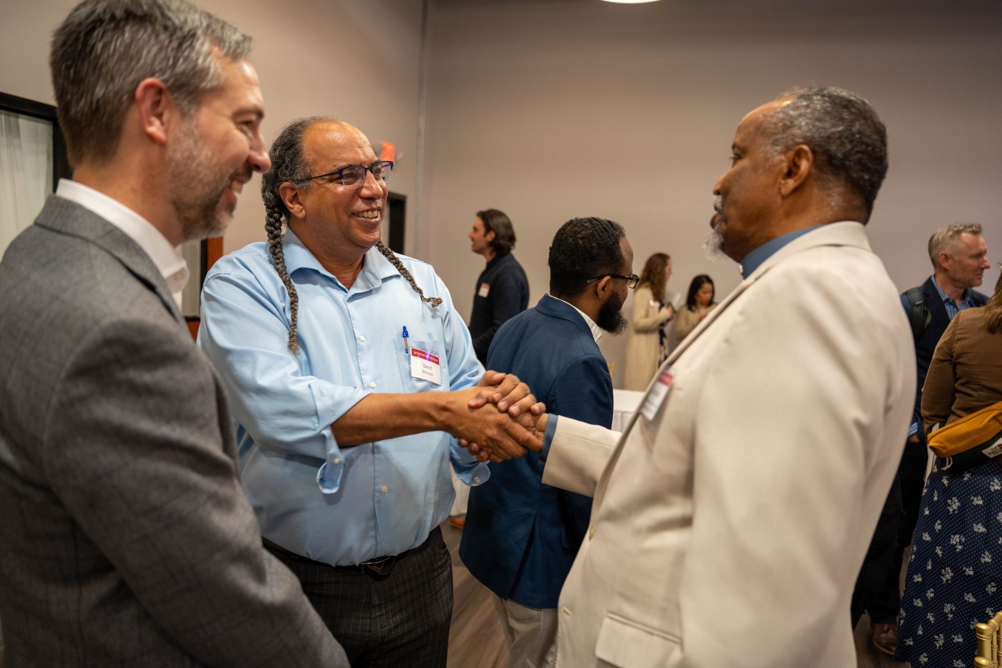 In a room of mingling people, a multiracial group of three men shake hands. Left to right: former Initiative Foundation President Matt Varilek. Vibrant & Equitable Communities Director David Nicholson, and Jama Alimed, an elder and leader in St. Cloud's Somali Community.