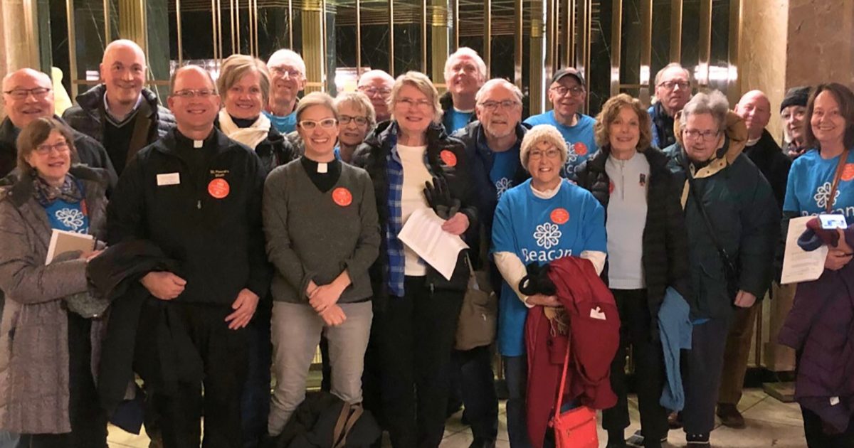 A group of people in a formal lobby. Some wear matching blue t-shirts with the Beacon Interfaith logo.