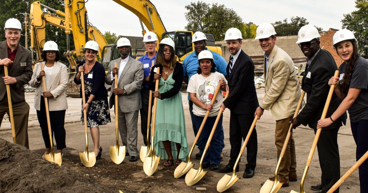 A group of people at a construction site wearing hard hats and holding shovels in a celebratory pose